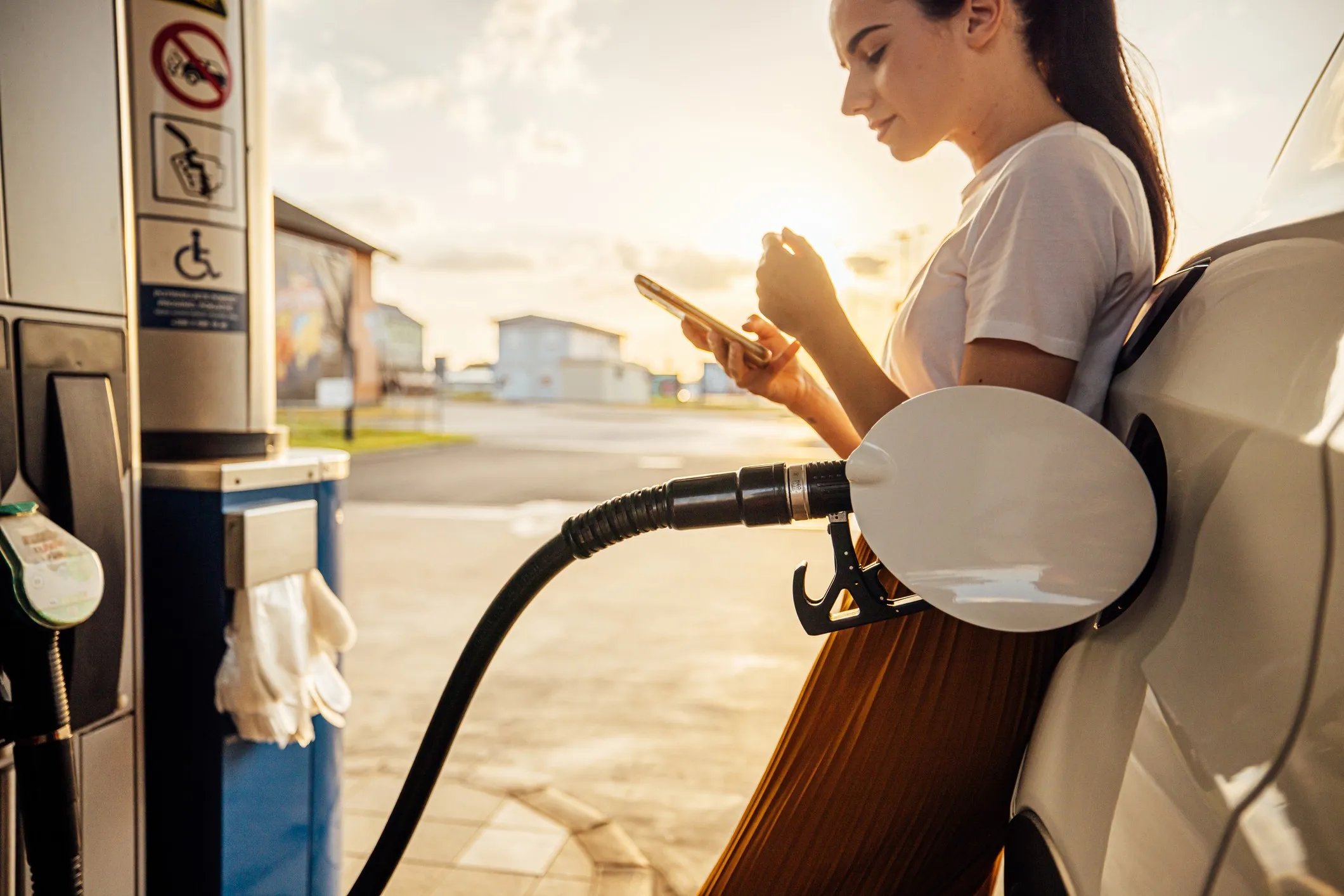 Young woman uses her smartphone to browse tips on how to save money on gas while refueling at the gas pump