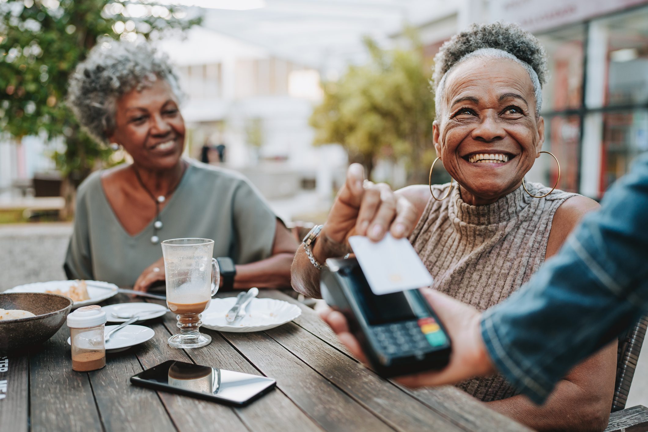 Two smiling women enjoy a meal at an outdoor cafe while one makes a contactless credit card payment, highlighting the importance of maintaining a good payment history.